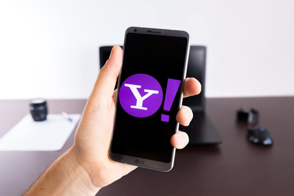 Logotipo do Yahoo é exibido na tela de um smartphone que por sua vez é segurado por uma mão ao fundo há uma mesa de escritório com um notebook e um mouse