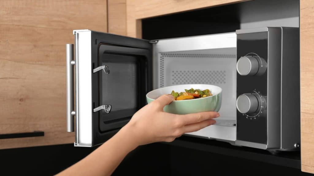 mãos de mulher colocando uma tigela com comida dentro do micro ondas na cozinha